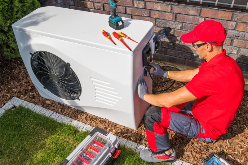4 Factors to Consider When Buying a Heat Pump. Professional Middle Aged HVAC Technician in Red Uniform Repairing Modern Heat Pump Unit.
