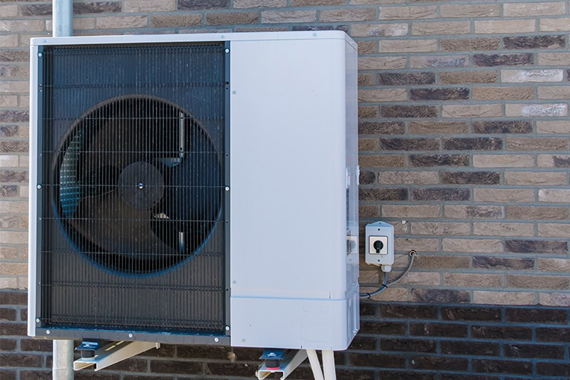 A large, modern heat pump or air conditioning condenser unit installed outdoors against a brown brick wall.