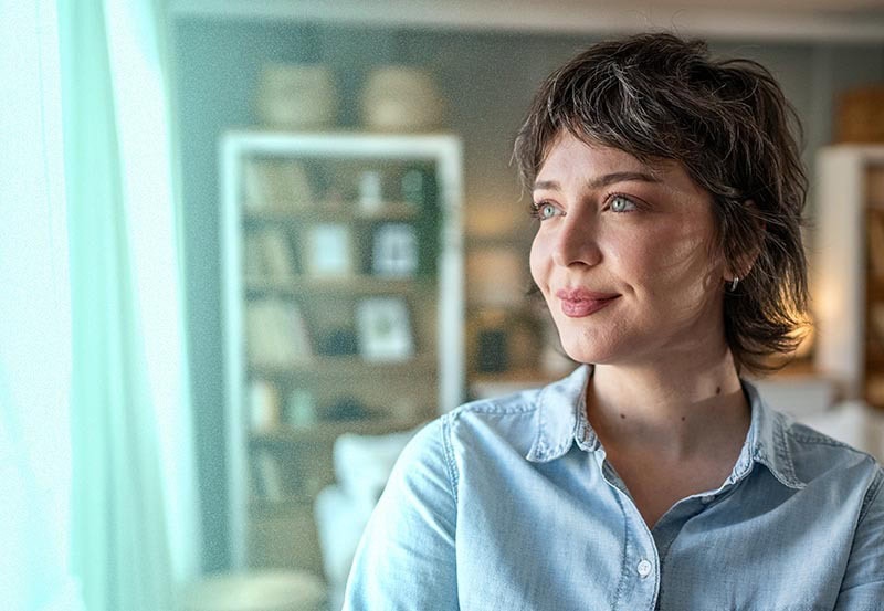 An image of a woman with short, wavy dark hair and light eyes, wearing a light blue denim shirt. She has a subtle smile and is looking off camera toward a window with soft, light green curtains, with a blurred living room interior and bookshelf visible in the background.